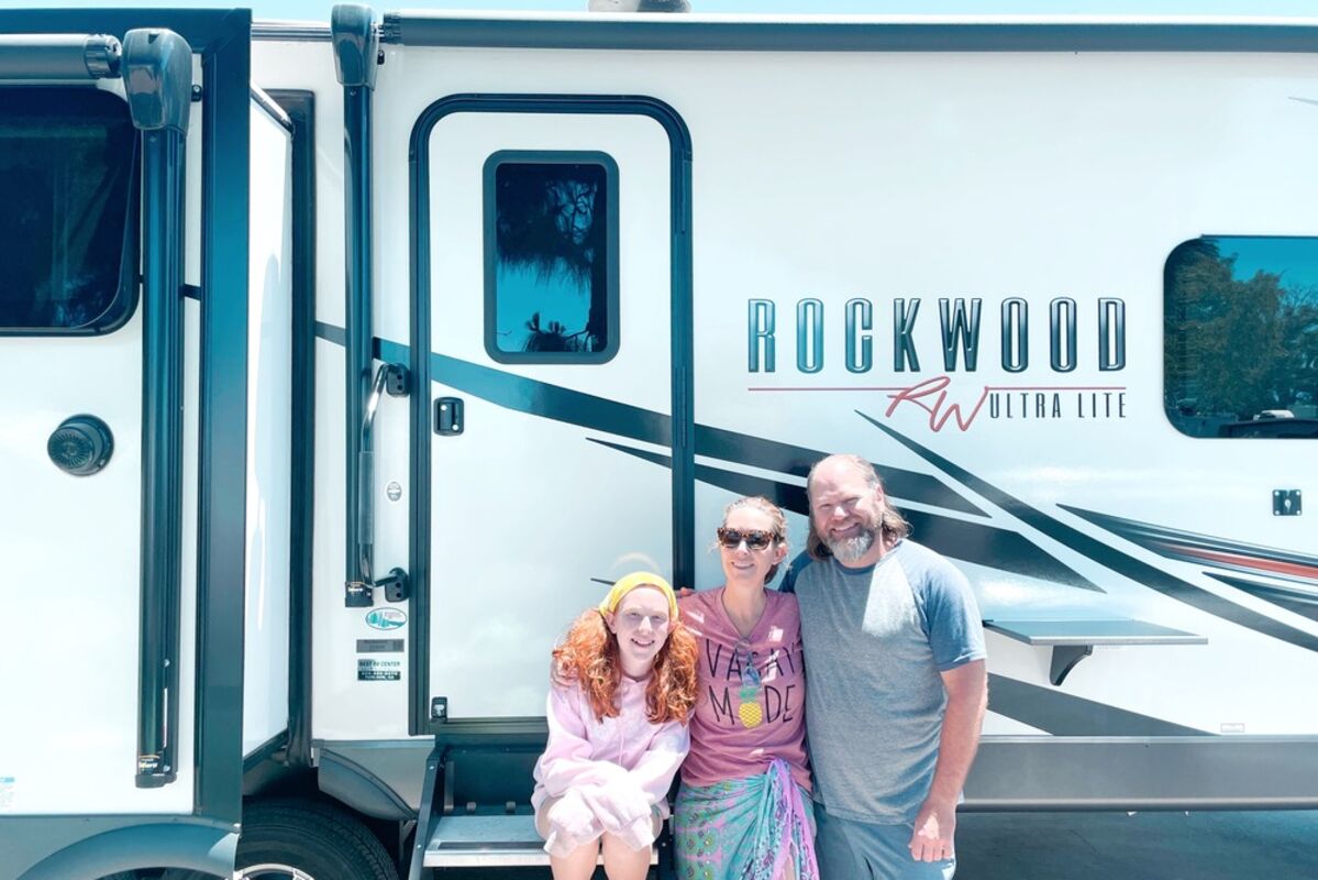 A family of 3 standing in front of an RV that says Rockwood on it.