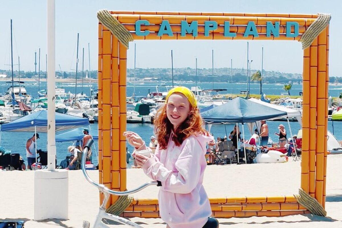 Girl on bike eating ice cream at the beach with a Campland sign.